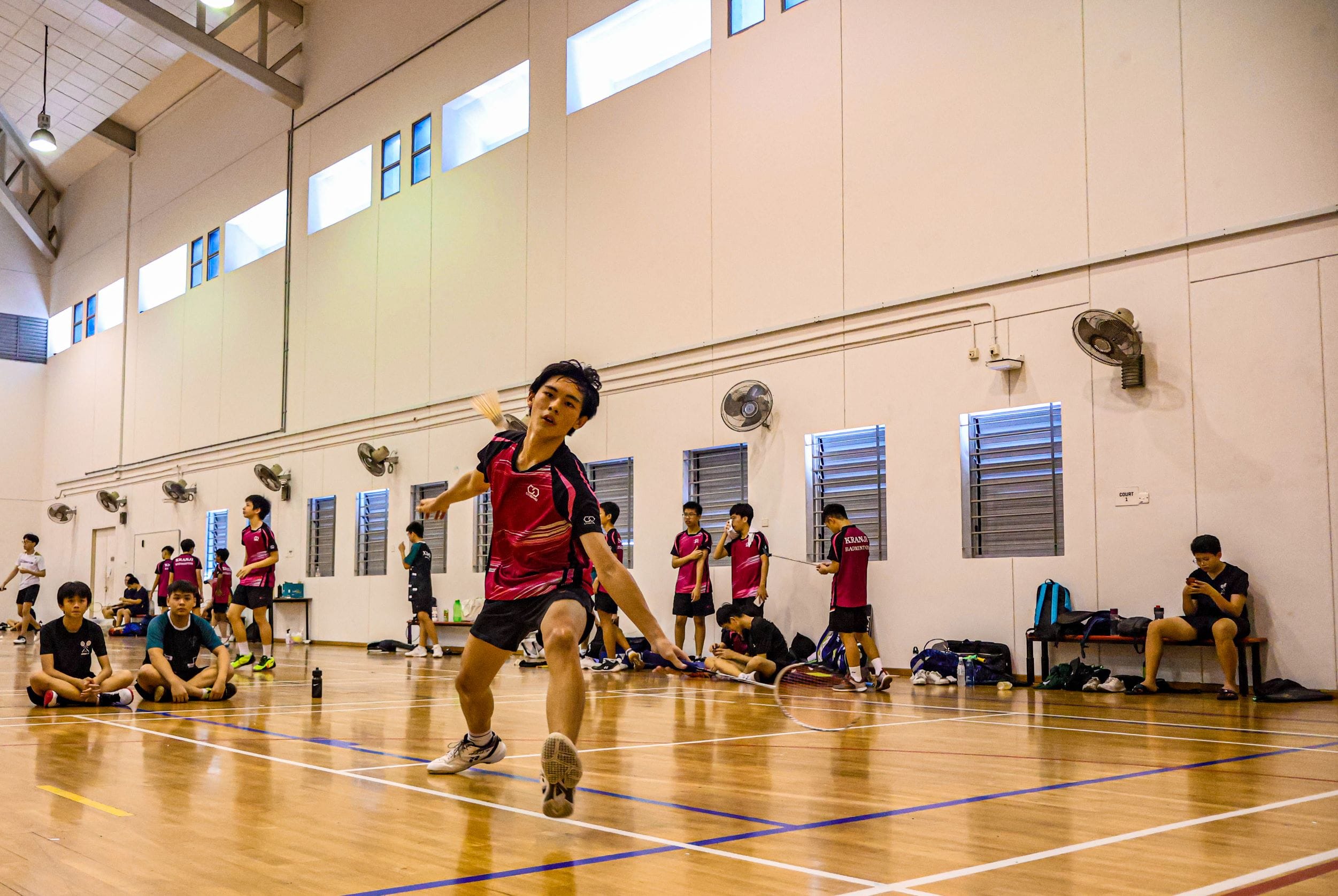 A young player in a pink jersey swings a badminton racket mid-lunge on an indoor court with teammates nearby.